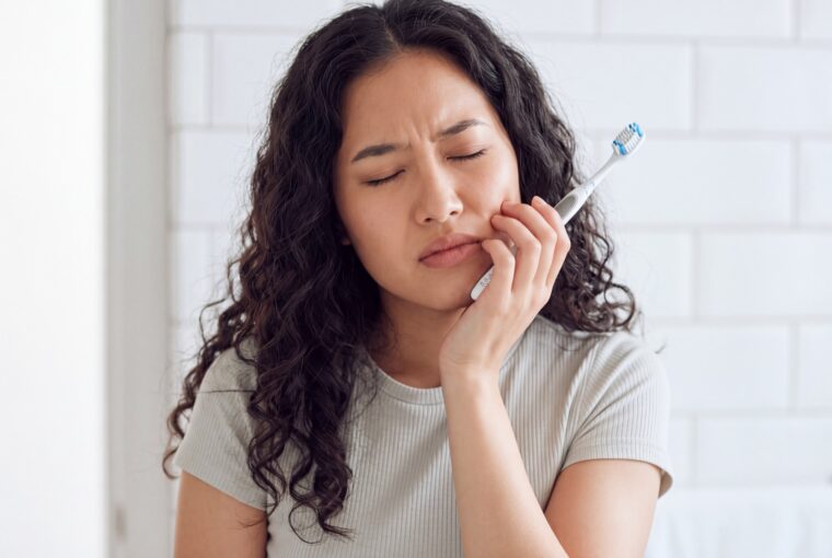Woman holding her jaw in discomfort while brushing her teeth