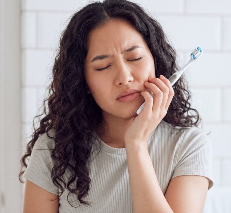 Woman holding her jaw in discomfort while brushing her teeth